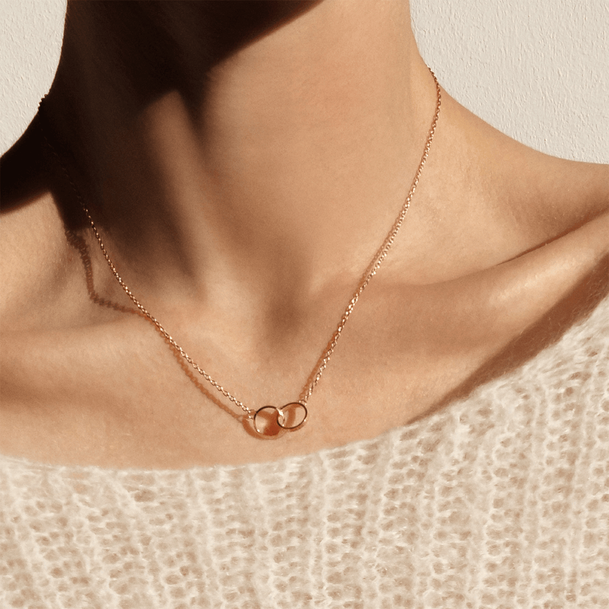 The Elan Unity Necklace Rose Gold worn at the collarbone, featuring two interlocking rose gold rings on a fine chain, styled with a soft knit top for an elegant everyday look.
