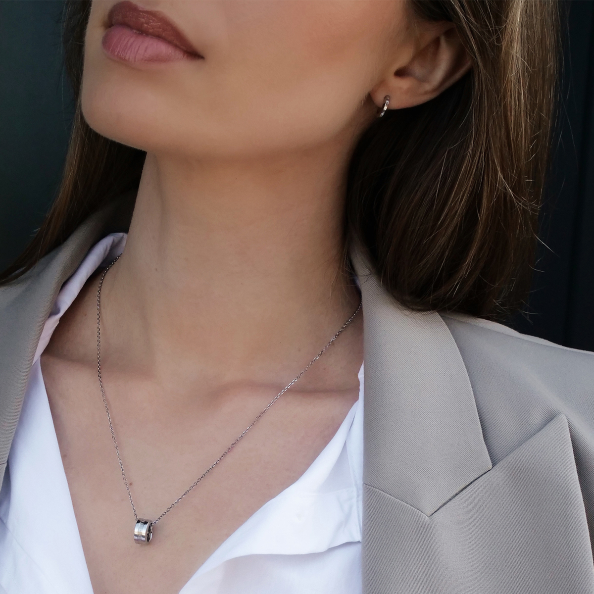 A woman wears the Elan Earrings Silver, styled with a neutral blazer and white shirt, highlighting the minimalist silver huggie earrings as a refined accessory for everyday or business wear.