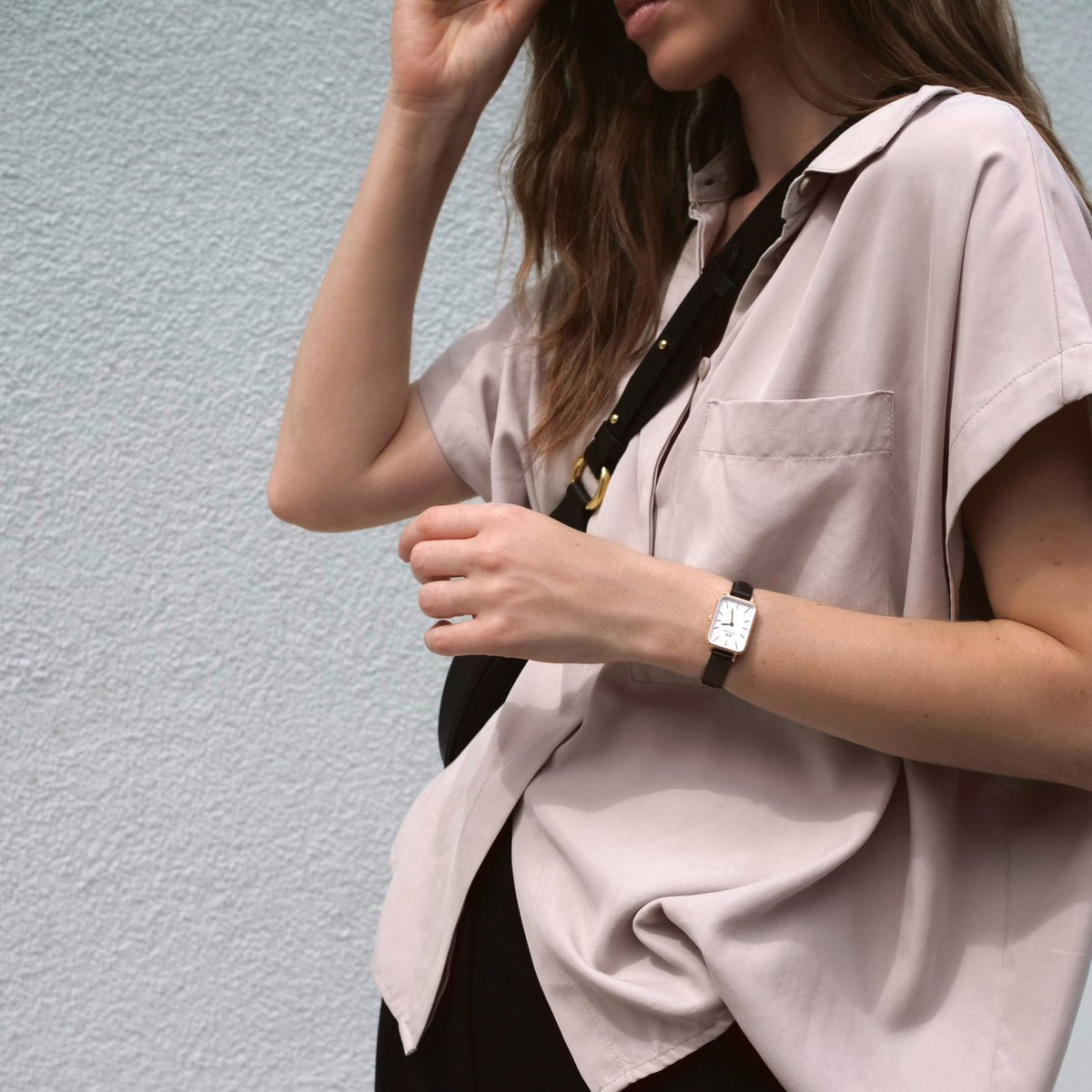 A woman wearing the Quadro Pressed Sheffield Rose Gold watch with a rectangular white dial, rose gold detailing, and a slim black leather strap, styled casually with a light short-sleeve shirt and crossbody bag.