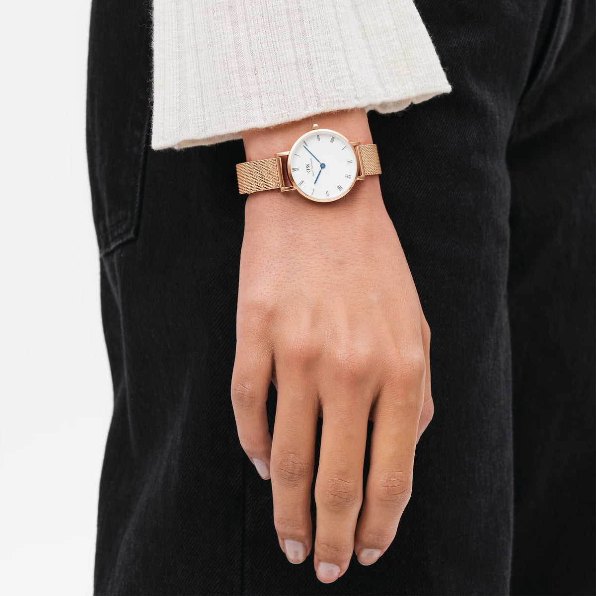 Petite Roman Numerals Melrose ladies watch with a rose gold mesh strap and white Roman numeral dial, worn on the wrist with casual black trousers and a cream knit top.