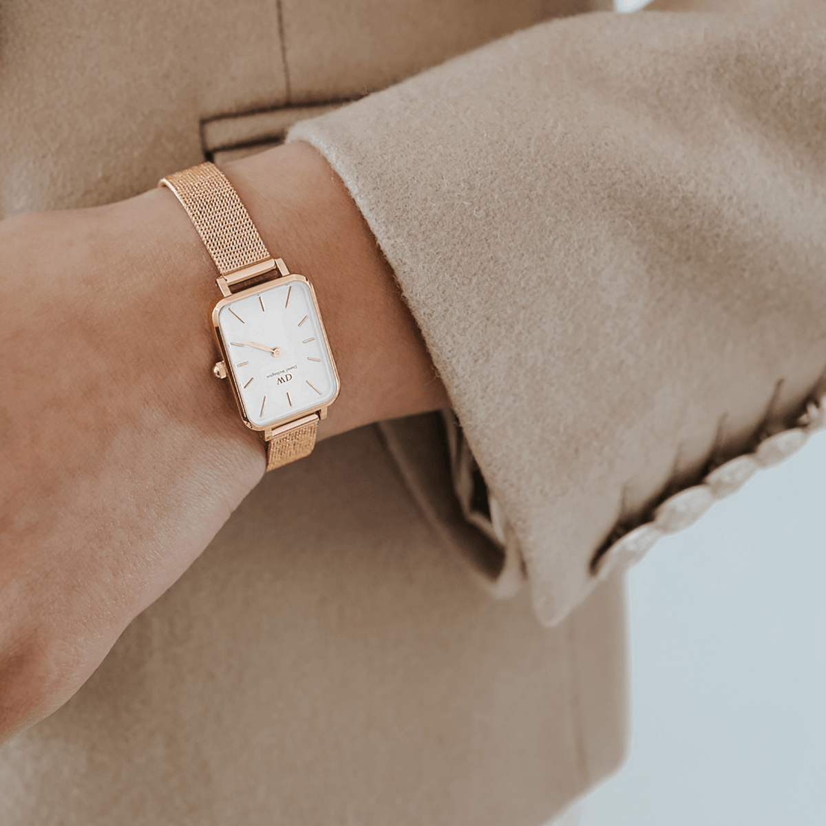 Quadro Pressed Melrose Rose Gold ladies watch with a rectangular case, white minimalist dial, and rose gold mesh strap worn on a wrist, styled with a beige tailored coat.