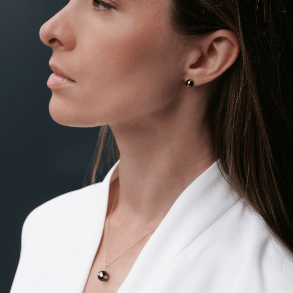 A woman wearing Aspiration Earrings Rose Gold Black, featuring polished black ceramic spheres with a rose gold plated band, styled with a matching pendant and a white blazer for a sophisticated, modern look.
