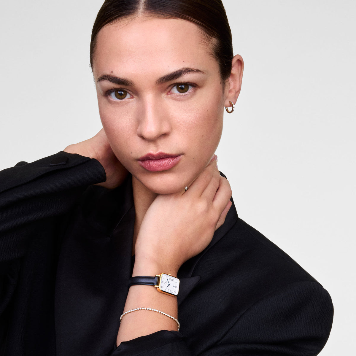 Quadro Roman Numerals Date Sheffield Gold ladies watch with a rectangular gold case and black leather strap, worn with a black blazer and a delicate bracelet for a refined, modern look.