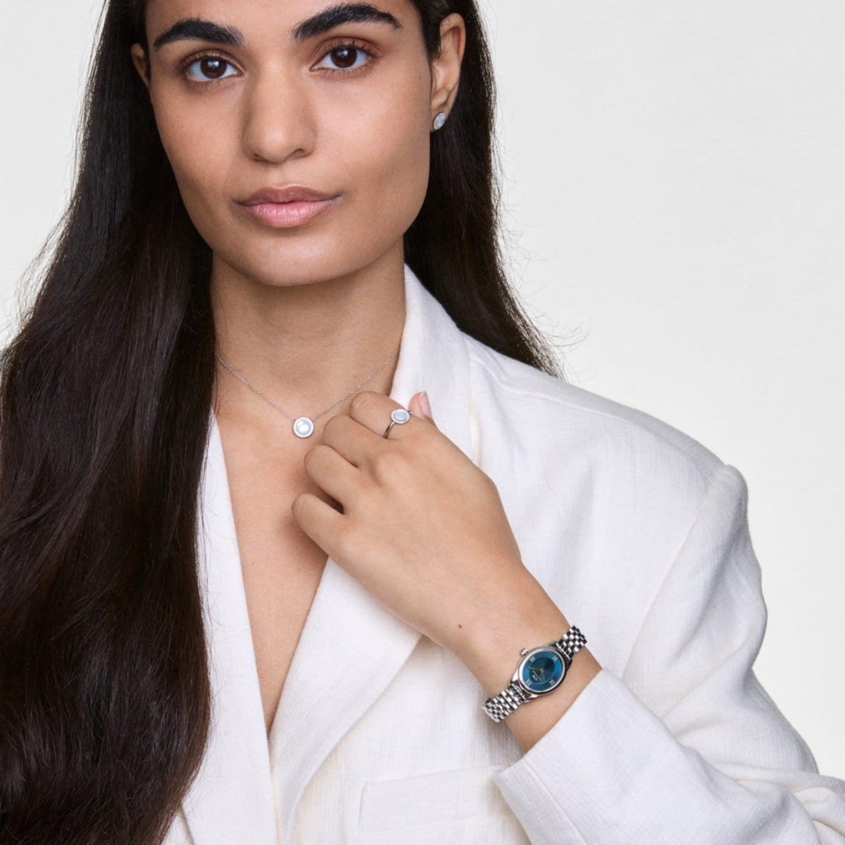 A woman wearing the Ophelia Mini Arctic Silver watch, featuring a polished oval silver case, blue sunray dial, and slim 5-link bracelet, styled with a white blazer and silver jewellery for a refined, modern look.
