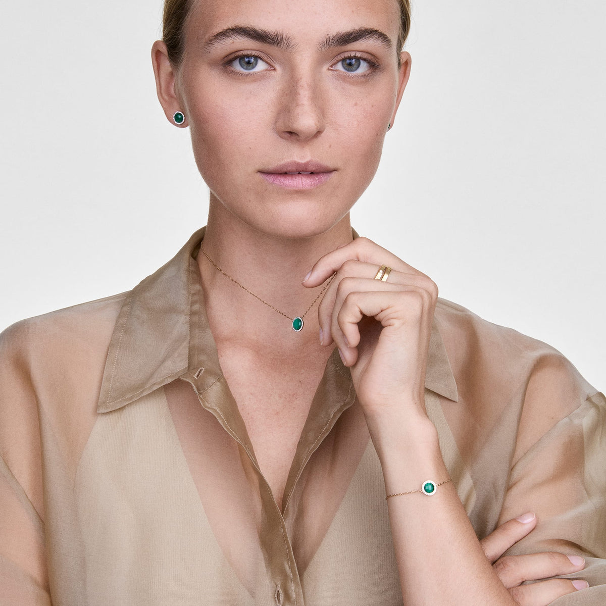A woman wearing the Audrey Necklace Malachite Gold, styled with a sheer beige blouse, shows the gold chain and green malachite pendant with cubic zirconia accents, paired with matching jewellery for a coordinated and elegant look.