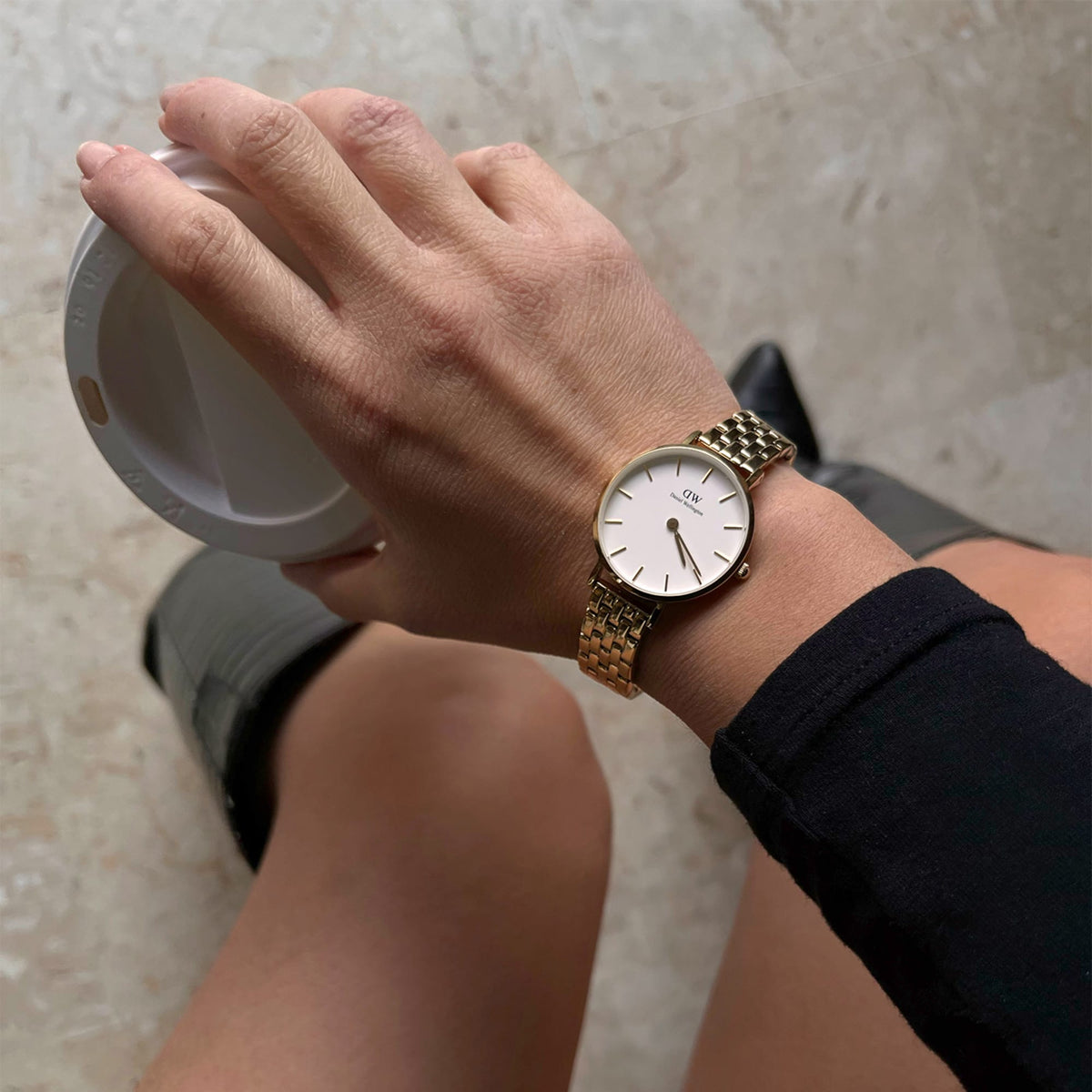 A woman wearing the Petite 5-Link Evergold gold watch with a minimalist white dial and 5-link bracelet, styled casually while holding a coffee cup, highlighting its elegant and versatile design for everyday wear.