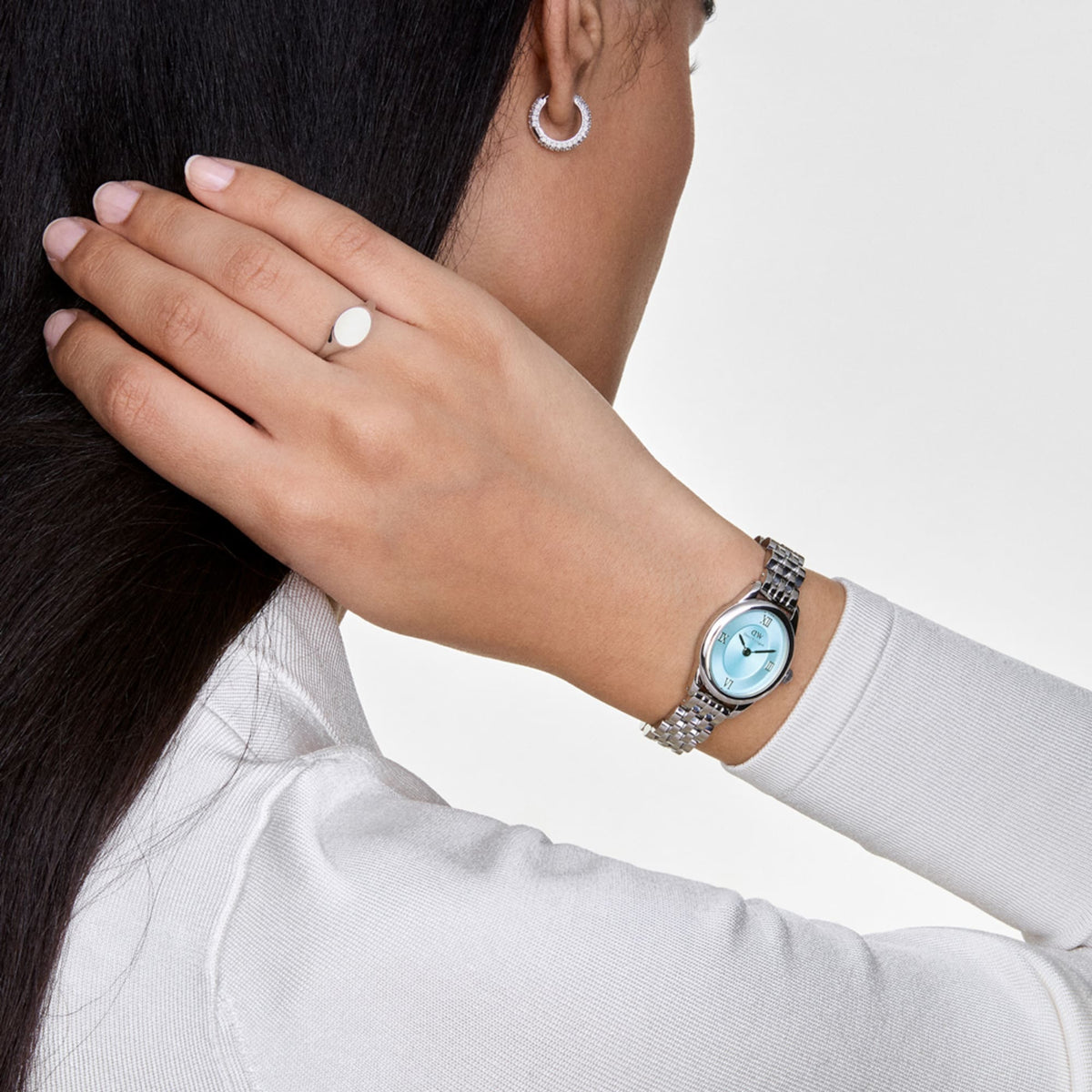 Classic Mini Signet Ring Silver worn on the finger, showcasing its minimalist oval design and polished stainless steel finish, paired with a silver watch and hoop earring for a modern, elegant look.