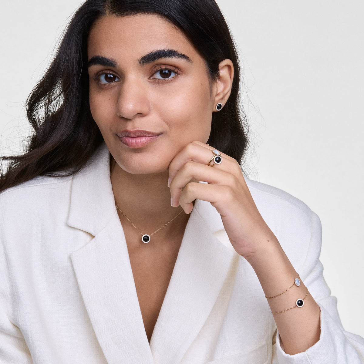 A woman wearing the Audrey Necklace Black Agathe Gold, featuring a gold chain and a black agate pendant with a halo of white cubic zirconia crystals, styled with matching jewellery and a cream blazer for a polished, modern look.