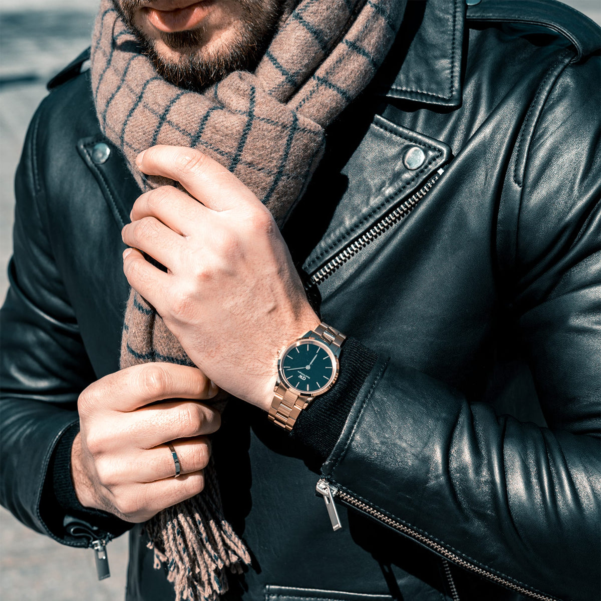 A man wearing the Daniel Wellington Iconic Link Black Rose Gold watch with a black dial and rose gold bracelet strap, styled with a black leather jacket and a checked brown scarf.