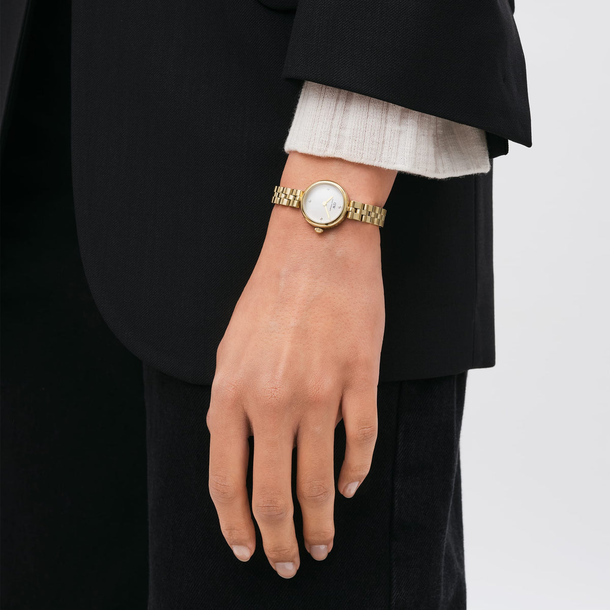 A woman wearing the Elan Lumine Gold watch with a slim gold bracelet and crystal-accented white dial, styled with a black blazer and cream knit top for a polished, everyday look.