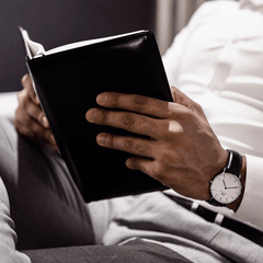 A person wearing the Classic Sheffield Silver watch with a black leather strap and minimalist silver case, reading a book while dressed in a white shirt and grey trousers in a relaxed indoor setting.
