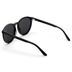 Arch Bio-Acetate Black sunglasses with round black frames, deep black lenses, and Daniel Wellington branding on the temple, shown from a rear angle to highlight the slim, lightweight bio-acetate arms.