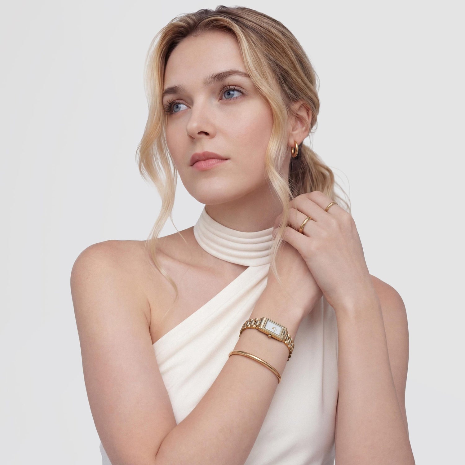 A woman wearing the Bound Mini 3-Link Gold watch with a rectangular gold case and 3-link bracelet, styled with gold jewellery and a white one-shoulder top for an elegant, modern look.