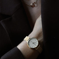 A woman wearing the Daniel Wellington Petite Evergold gold mesh strap watch with a minimalist eggshell white dial, styled with a dark blazer for a refined and understated look.