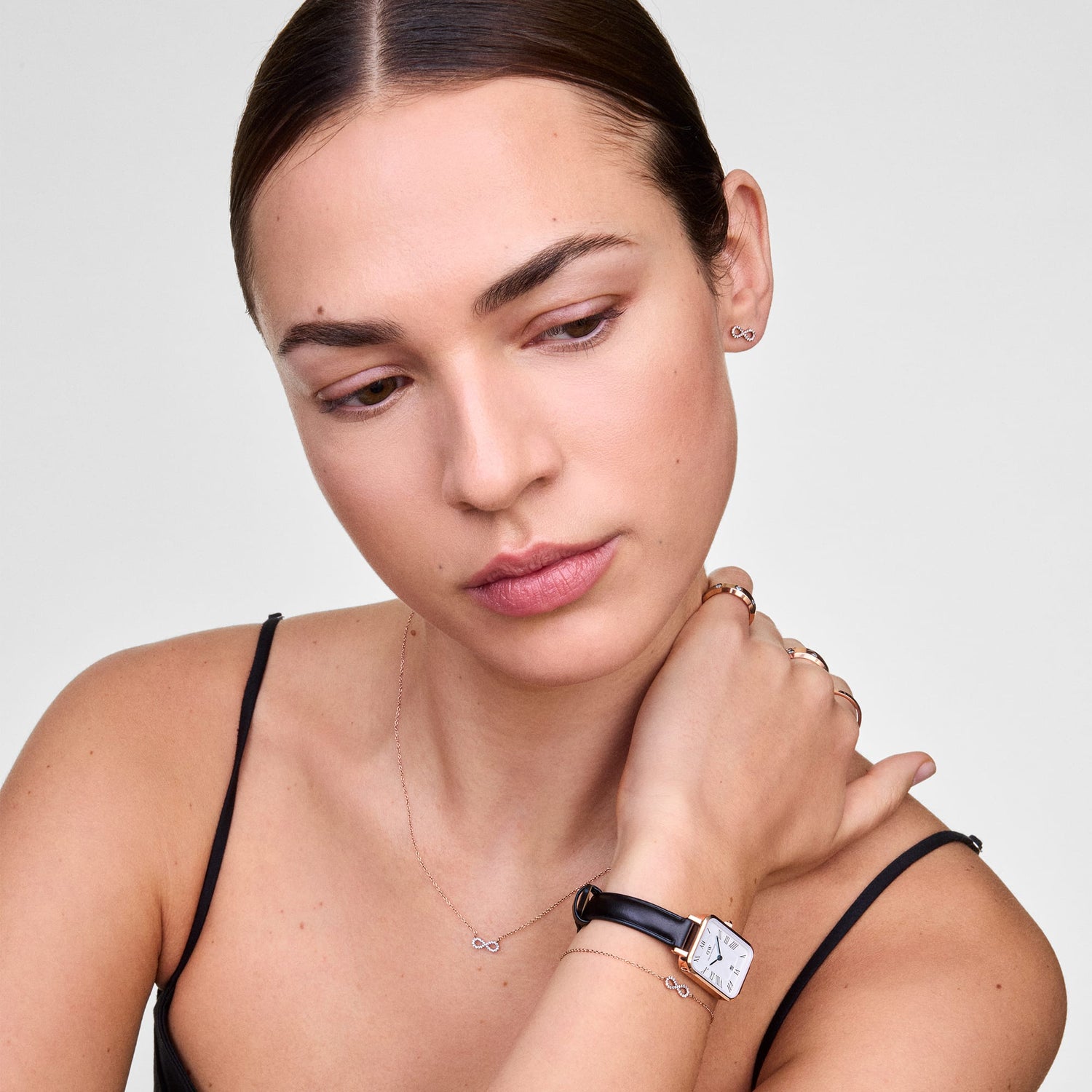 Quadro Roman Numerals Date Sheffield Rose Gold ladies watch worn on the wrist, featuring a rectangular rose gold case, white dial with Roman numerals, blue hands, date window, and a slim black leather strap styled with delicate jewellery.