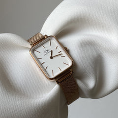 Quadro Pressed Melrose Rose Gold watch with a slim rectangular case, minimalist white dial, and rose gold mesh strap displayed elegantly on a textured white fabric background.