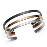 Three minimalist cuff bracelets in black, rose gold, and silver stainless steel, each engraved with Daniel Wellington branding and featuring a sleek, adjustable design suitable for layering or wearing alone.