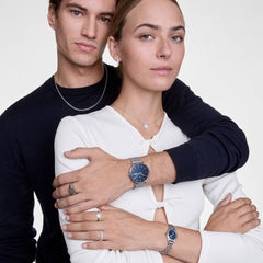 Couple wearing the Classic Day & Ophelia Mini Arctic Silver Set, featuring a men’s silver mesh watch with a blue dial and day display, and a women’s oval silver wristwatch with a blue dial and Roman numerals, styled for a modern and elegant look.