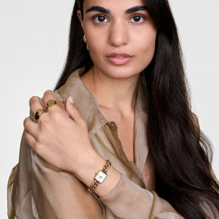 A woman wearing the Jolie Chain Gold watch, featuring a chunky gold chain bracelet and square white dial, styled with gold rings and a sheer beige blouse for a chic, modern look.