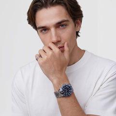 A man wearing the Daniel Wellington Iconic GMT Link Black Blue Ceramic Bezel Silver watch, styled with a silver stainless steel bracelet and a black and blue ceramic bezel, paired with a casual white T-shirt.
