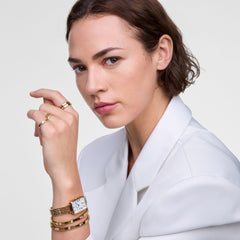 Quadro Roman Numerals Date 5-link Gold ladies watch styled on a wrist with gold bangles and rings, paired with a white blazer for a polished, modern look.