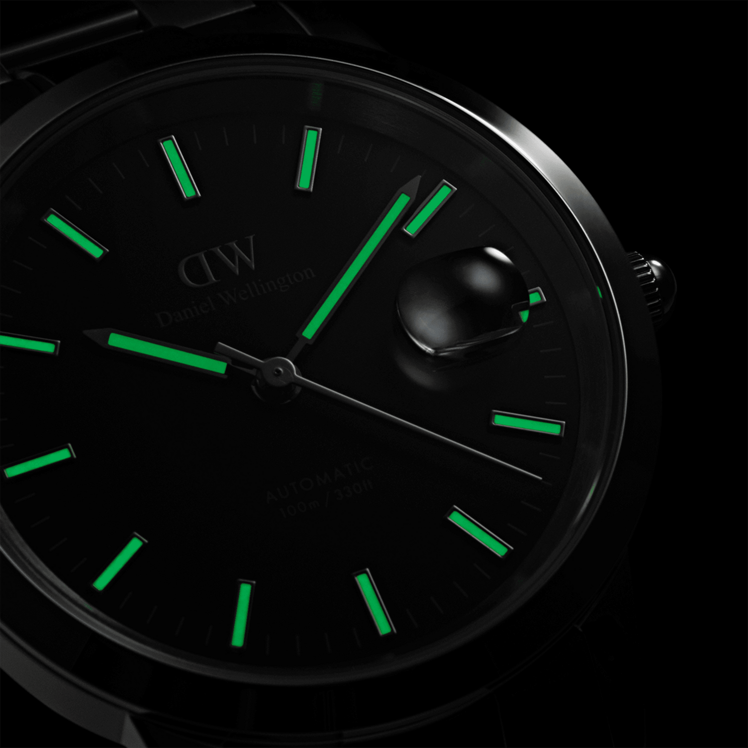 Daniel Wellington Iconic Link Automatic watch with luminous green markers and hands on a black dial, magnified date window, and polished stainless steel case, shown glowing in low light.