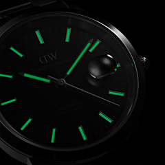 Daniel Wellington Iconic Link Automatic watch with luminous green markers and hands on a black dial, magnified date window, and polished stainless steel case, shown glowing in low light.