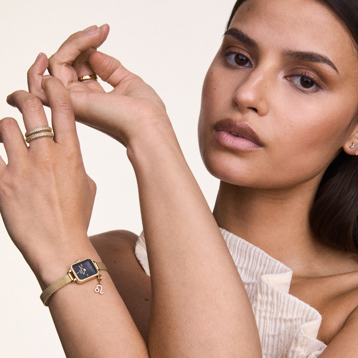 A woman wearing the Daniel Wellington Quadro Mini Crystal Zodiac Evergold Set, showcasing the gold mesh strap, crystal-framed rectangular case, black mother of pearl dial, and a sparkling zodiac charm, styled with gold rings for a coordinated and elegant look.