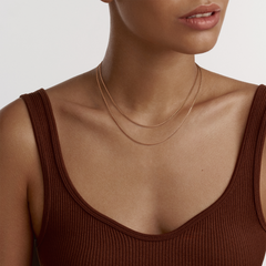 A woman wearing the Box Chain Necklace RG in rose gold, styled as a layered necklace that sits elegantly on the collarbone with a minimalist design, paired with a brown ribbed top.