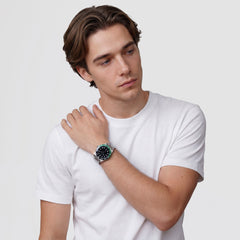 A man wearing the Daniel Wellington Iconic GMT Link Black Green Ceramic Bezel Silver watch, styled with a casual white t-shirt, showing the black dial, green and black ceramic bezel, and silver stainless steel bracelet on his wrist.