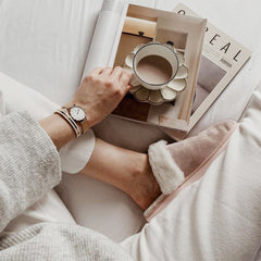 A person relaxing in cosy loungewear and slippers, wearing the Emalie Bracelet Satin White Rose Gold alongside a classic watch, enjoying a hot drink and reading a magazine.
