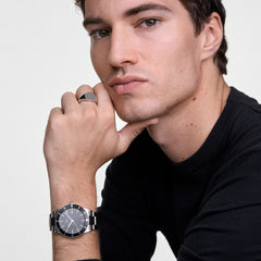 A man wearing the Daniel Wellington Iconic Paradigma Black & Classic Signet Ring Set, featuring a polished silver signet ring with a black face and a stainless steel watch with a black dial, styled with a black jumper for a modern, refined look.