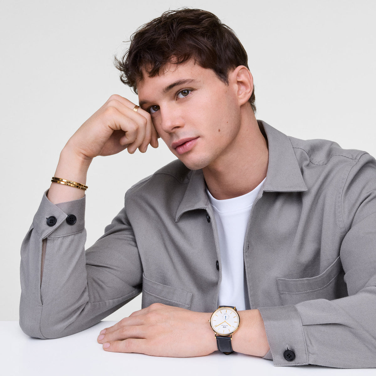 A man wearing the Classic Multi-Eye Sheffield Off-white Sunray Gold watch with a gold case, off-white dial, blue-tipped sub-second hand, and black leather strap, styled with a grey shirt and gold bracelet for a smart-casual look.