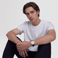 A man wearing the Classic Roman Numerals Date Black Croc Belly Rose Gold watch, styled with a white t-shirt and black trousers, showing the rose gold case, white dial with Roman numerals, blue hands, and black croc-embossed leather strap.