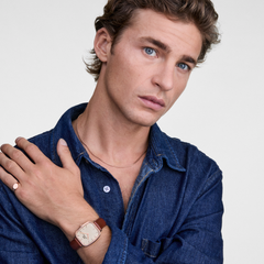 A man wearing a denim shirt displays the Marlon & Classic Mini Signet Ring Rose Gold Set, featuring a rose gold Daniel Wellington watch with a brown leather strap and a polished rose gold signet ring on his finger.