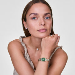 A woman wearing the Ophelia Mini Emerald Gold watch, featuring a slim gold beans link bracelet and an oval emerald green sunray dial, styled with a neutral sleeveless top and a delicate pendant necklace.