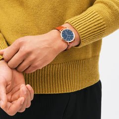 Classic Multi-Eye St Mawes Arctic Rose Gold men's watch with a blue chronograph dial and brown leather strap, worn on the wrist with a mustard knit jersey and black trousers.
