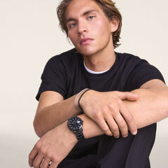 A man wearing the Daniel Wellington Iconic Link Automatic Black watch, featuring a black stainless steel link bracelet and a luminous black dial, styled with a casual black t-shirt for a modern, versatile look.