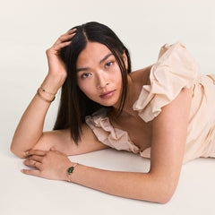 Petite Mini Emerald Melrose watch with emerald green dial and rose gold mesh strap worn by a woman in a light ruffled dress, styled with matching rose gold jewellery in a relaxed pose.
