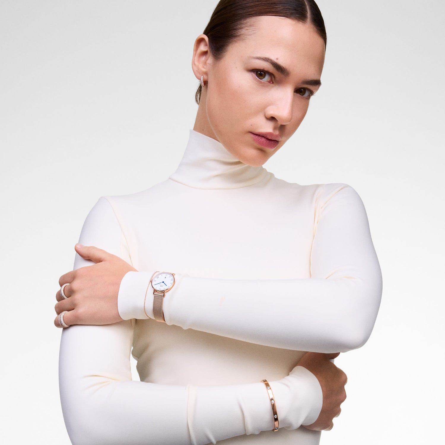 A woman wearing the Petite Roman Numerals Date Rose Gold watch with a slim rose gold mesh strap, white dial, and Roman numerals, styled with a matching rose gold bracelet and a cream long-sleeve top.
