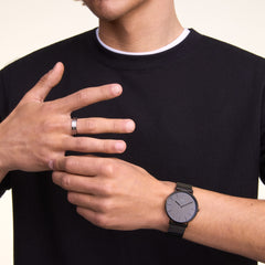 A man wearing the Classic Piano Link Onyx Unitone Black watch by Daniel Wellington, featuring a brushed black dial and geometric stainless steel bracelet, styled with a casual black T-shirt for a modern, understated look.