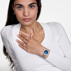 A woman wearing the Daniel Wellington Crystalline Bezel Sterling Blue watch with a blue mother of pearl dial, crystal-set bezel, and silver mesh strap, styled with matching silver jewellery and a white top.