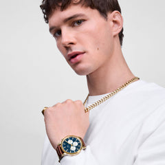 A man wearing the Iconic Chronograph St Mawes Arctic Panda Gold watch, featuring a gold case, blue panda dial with white sub-dials, and a brown leather strap, styled with a gold chain and ring for a modern, polished look.