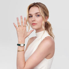 A woman wearing the Daniel Wellington Ophelia Mini Green Lizard White Guilloché Gold watch on her wrist, styled with gold jewellery and a sleeveless white top, highlighting the elegant green lizard-embossed leather strap and petite gold oval case.
