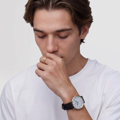 A man wearing the Daniel Wellington Classic Roman Numerals Date Black Croc Belly Gold watch, featuring a gold case, white dial with Roman numerals, blue hands, and a black croc-embossed leather strap, styled with a casual white T-shirt.