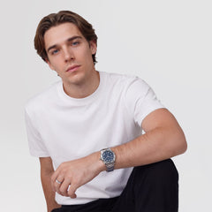 A man wearing the Daniel Wellington Iconic GMT Link Brushed Bezel Silver watch with a blue dial and stainless steel bracelet, styled casually with a white T-shirt and black trousers.