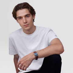A man wearing the Daniel Wellington Bound Light Brown Arctic Guilloché Silver watch, featuring a rectangular silver case, blue textured dial, and light brown leather strap, styled casually with a plain white T-shirt.