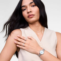 A woman wears the Petite Roman Numerals Date Silver watch with a slim silver mesh strap, blue hands, and Roman numerals, styled with minimalist jewellery and a neutral sleeveless top for a modern, elegant look.