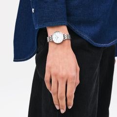 Petite Roman Numerals 5-Link Silver watch with a white dial, Roman numerals, and blue steel hands, worn on the wrist with a polished five-link stainless steel bracelet, styled casually with a denim shirt and black trousers.
