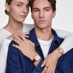 A couple wearing the Marlon & Jolie Chain Rose Gold Set by Daniel Wellington, featuring a men’s cushion-shaped rose gold watch with a brown leather strap and a women’s petite rectangular watch with a rose gold chain bracelet, styled with casual denim and white tops.